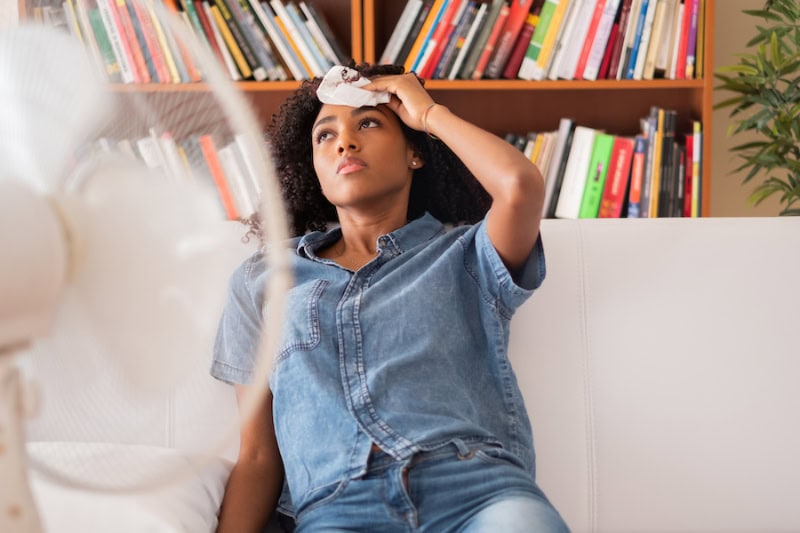 Woman Sitting in front of Fan Dabbing Her Forehead from Sweat.
