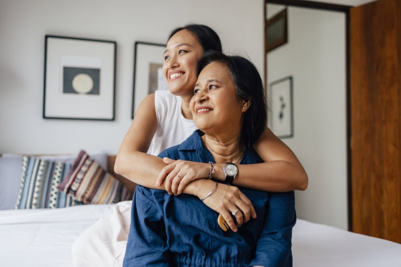 Standing daughter with her arms wrapped around her sitting mother.