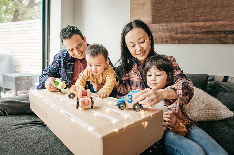 A cardboard box decorated with taped lanes and warm fairy lights serves as a homemade race track for numbered wooden toy cars.