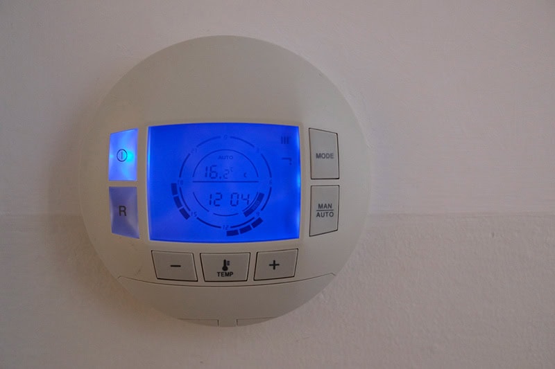 How to Set Your Programmable Thermostat. Photo of a digital thermostat mounted on the wall with a blue screen.